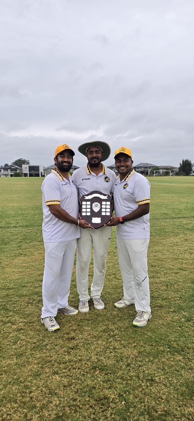 Three GT Comrades teammates stand on a cricket field holding a championship shield